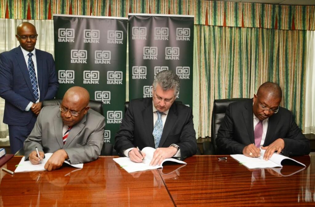 Three men seated at a boardroom table signing documents, with Co-operative Bank banners behind them and a fourth man standing.
