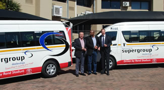 Three men standing between two Super Group branded minibuses outside a building.