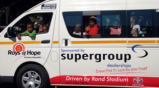 Children looking out of the windows of a minibus signwritten "Rays of Hope, sponsored by Super Group dealerships".