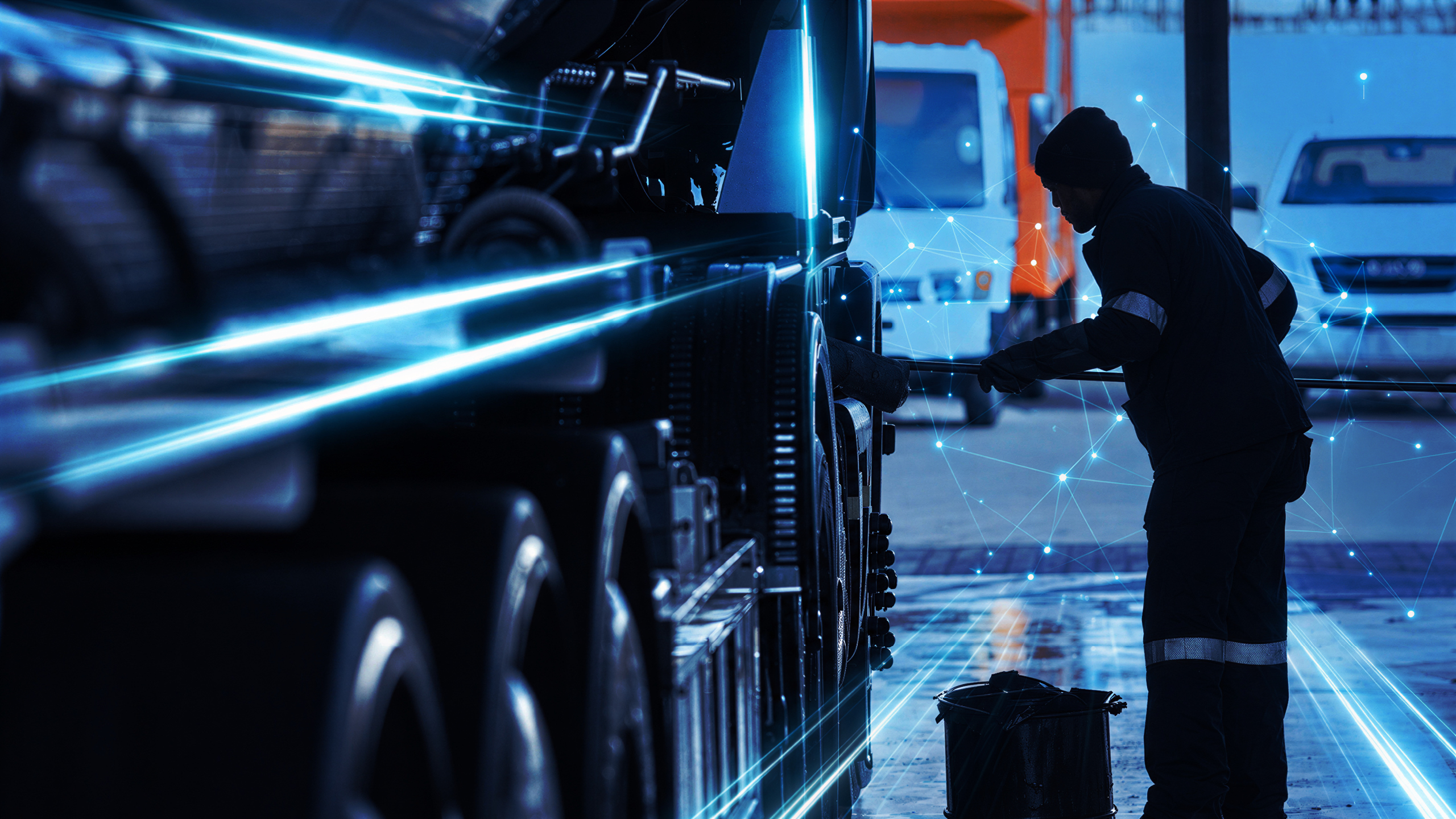 A worker in reflective-striped overalls inspects the wheels of a truck in a depot at night, lines of light running along the trailer.